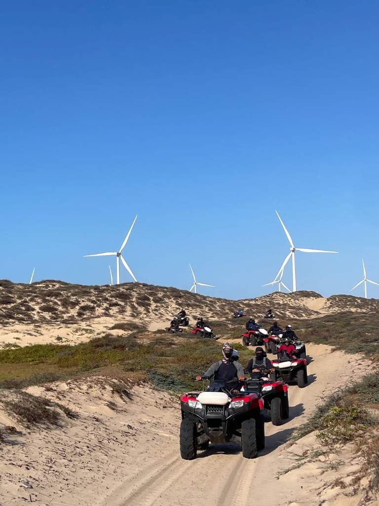 Comboio de quadriciclos em trilha de areia com aerogeradores ao fundo nos Lençóis Maranhenses