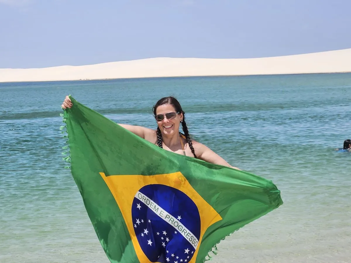 Visitante com bandeira do Brasil na beira de uma lagoa de águas cristalinas, com as dunas ao fundo, no passeio de Atins