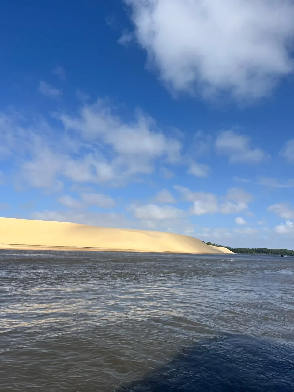 Duna dorada a orillas del río Preguiças, vista desde la lancha, bajo un cielo azul con nubes