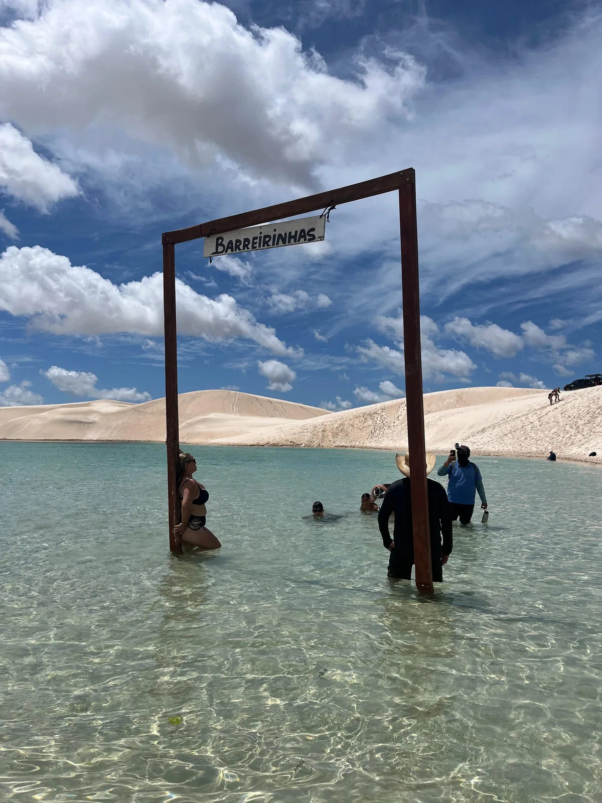 Visitantes na água cristalina da Lagoa Azul, sob o letreiro de Barreirinhas, com as dunas dos Lençóis ao fundo