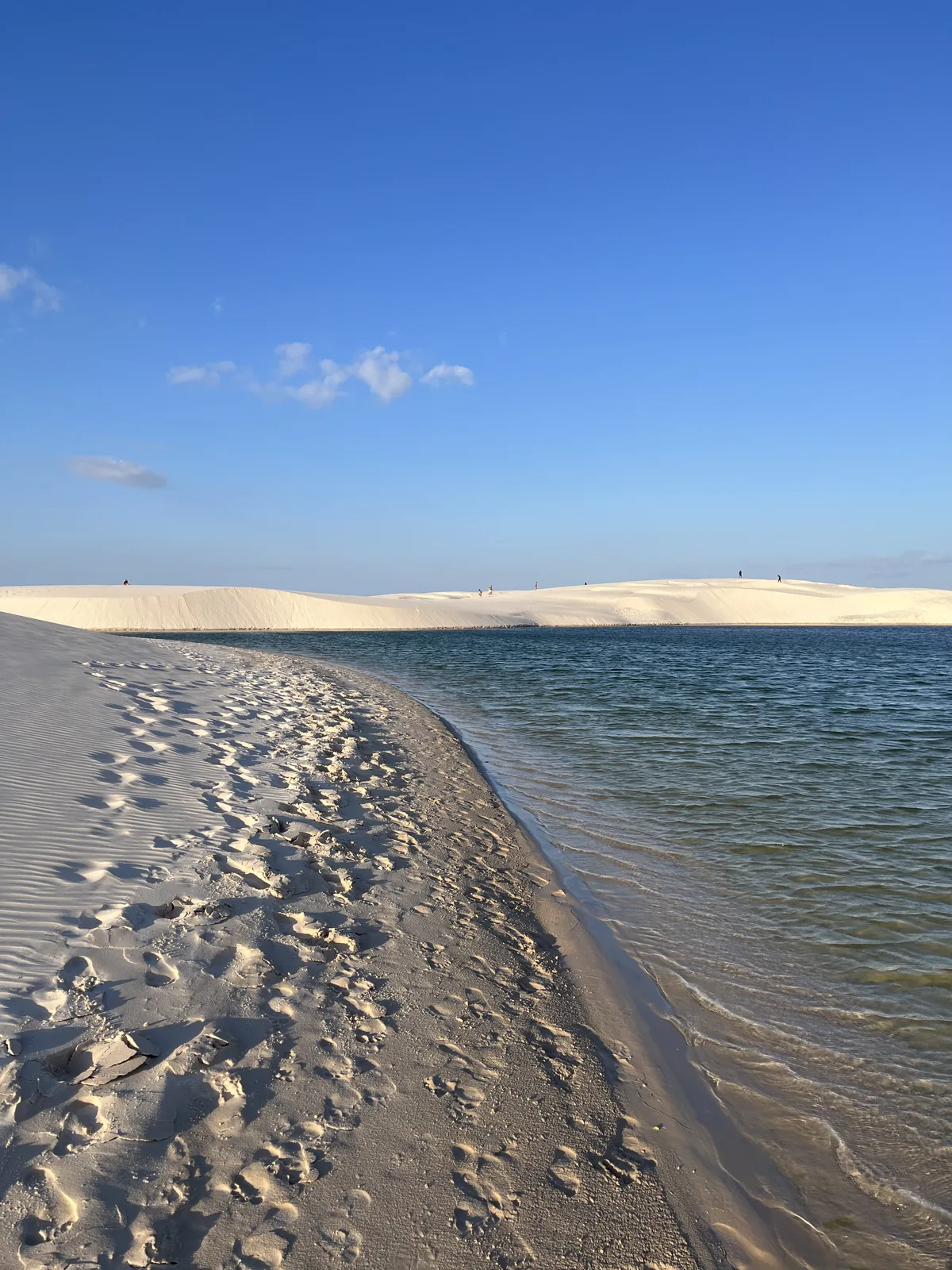 Strip of white sand along the edge of the lagoon, with the Lençóis dunes in the background