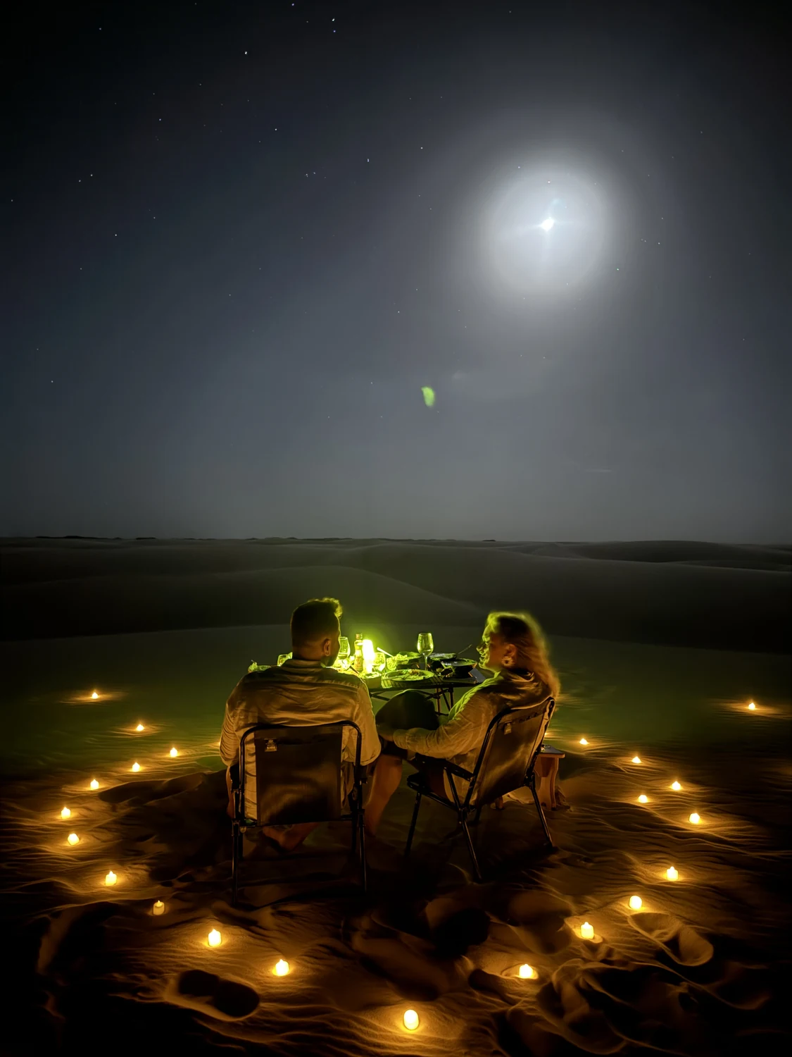 Casal jantando nas dunas sob a lua, cercado por velas acesas na areia