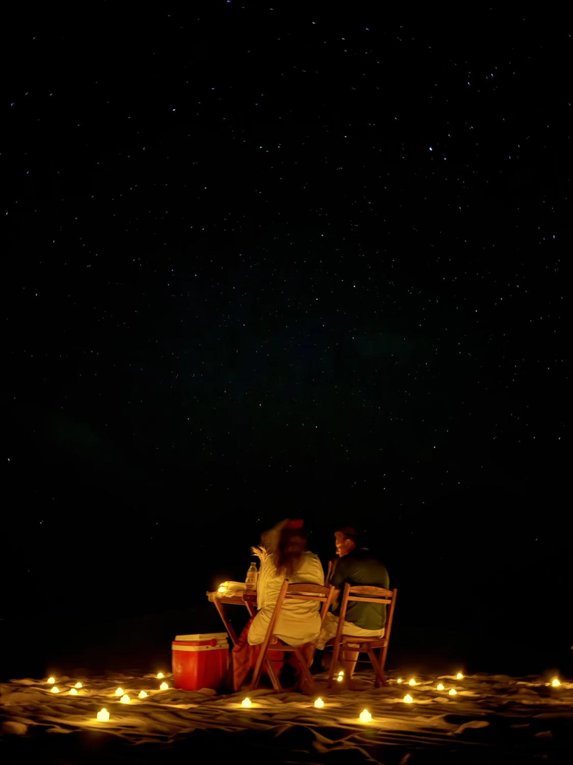 Casal à mesa nas dunas, com um círculo de velas acesas na areia e o céu estrelado