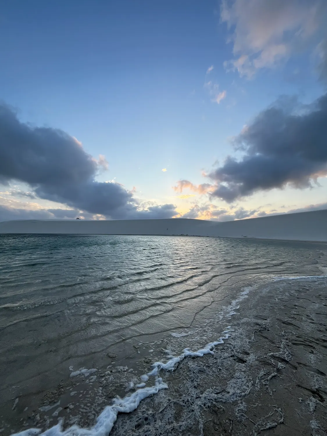 Lagoa dos Lençóis ao amanhecer, com a duna branca e as nuvens refletidas na água
