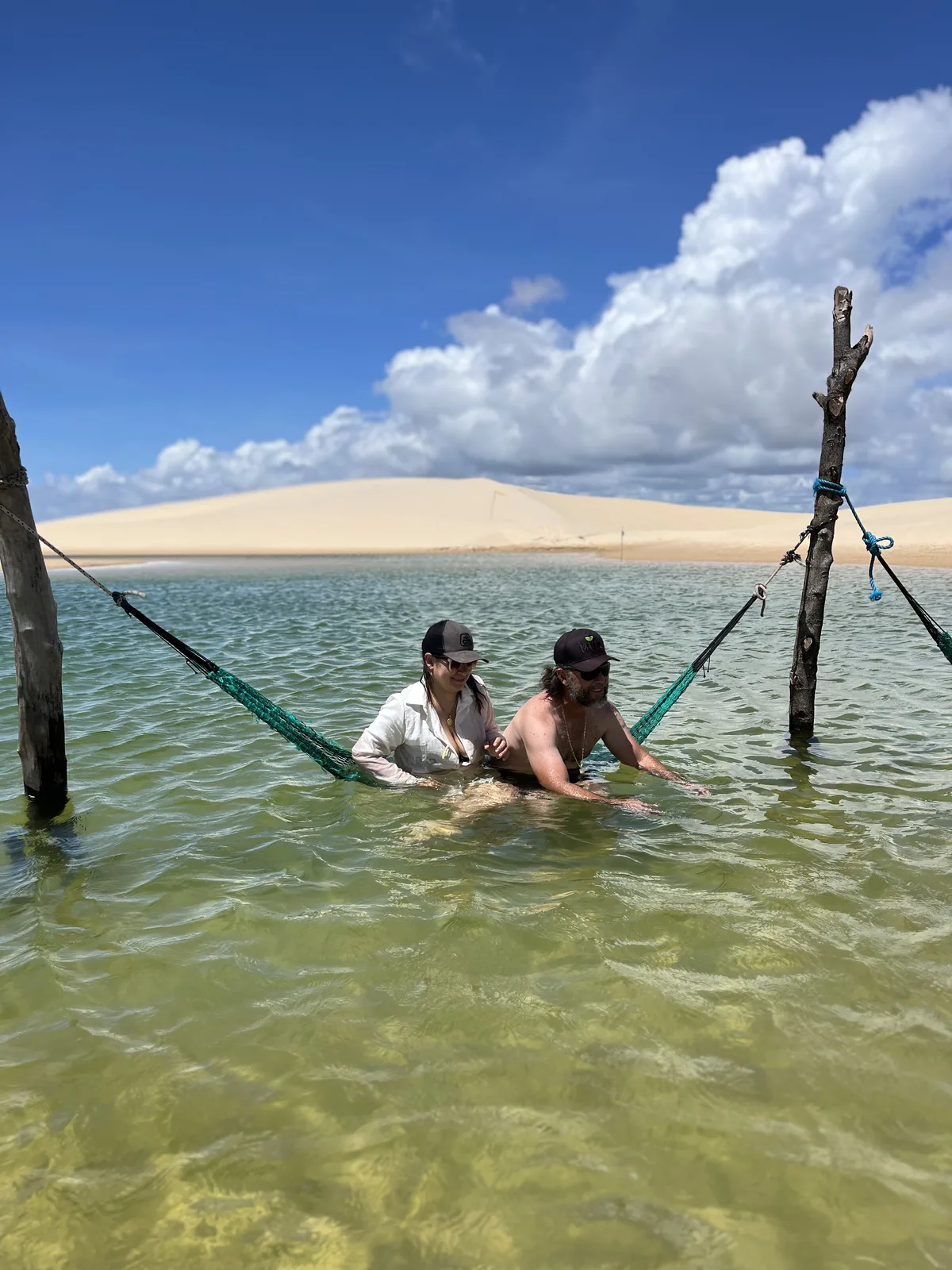 Casal em rede dentro da água na Lagoa do Alazão, com a duna ao fundo
