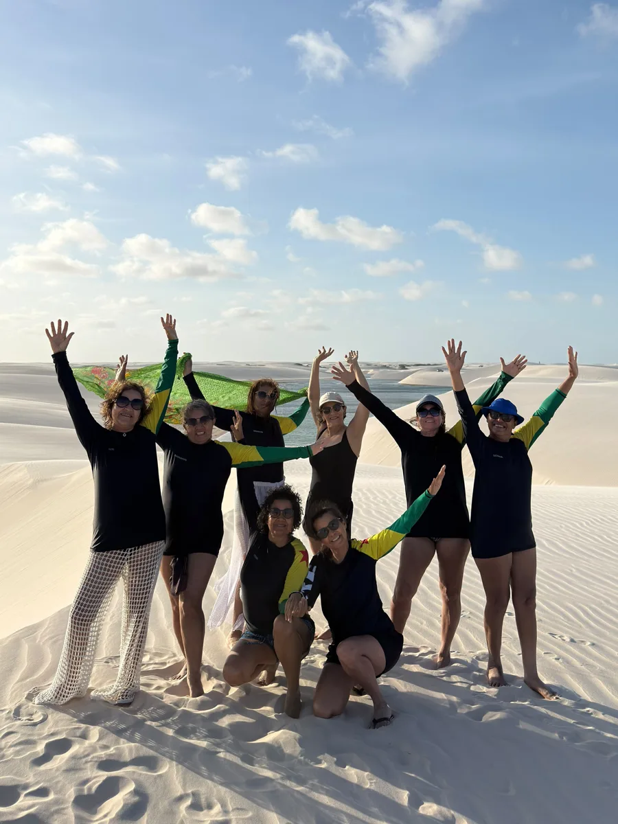 Grupo de viajantes comemorando de braços para o alto nas dunas dos Lençóis Maranhenses, com uma lagoa ao fundo