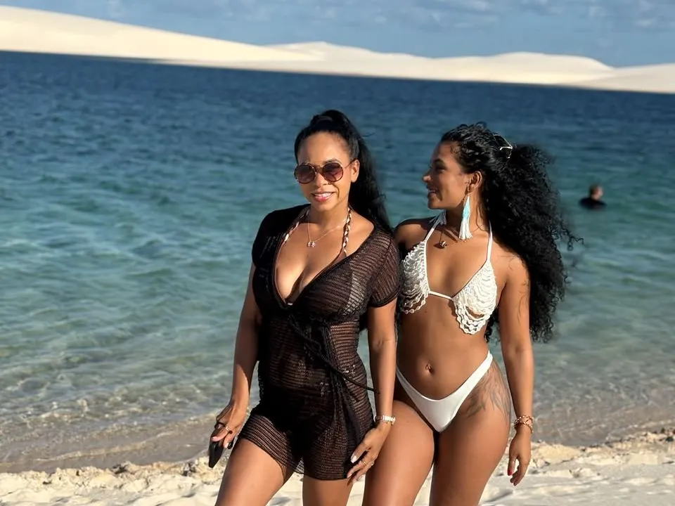 Duas visitantes na beira de uma lagoa de águas azuis, com as dunas ao fundo, no Circuito Santo Amaro