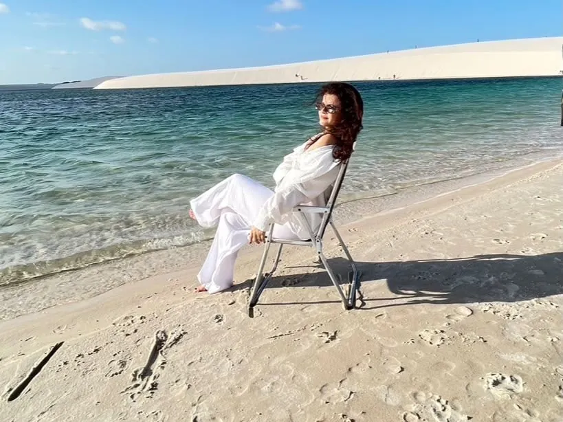 Visitante sentada em cadeira na beira de uma lagoa de águas cristalinas, com a duna ao fundo, no Circuito Santo Amaro