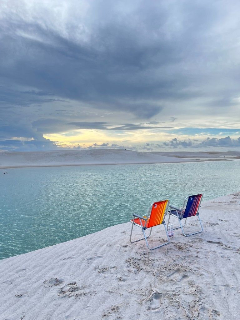Cadeiras de praia na borda de uma duna de areia branca, com lagoa turquesa e céu nublado nos Lençóis Maranhenses